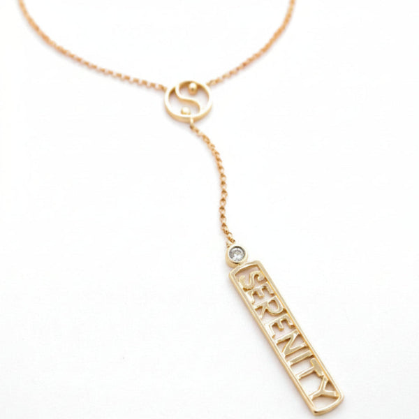 A solid gold Y-necklace featuring a small circular Yin Yang symbol with an long rectangular bar with the word 'SERENITY' and accented by a white diamond shown on a plain white background.